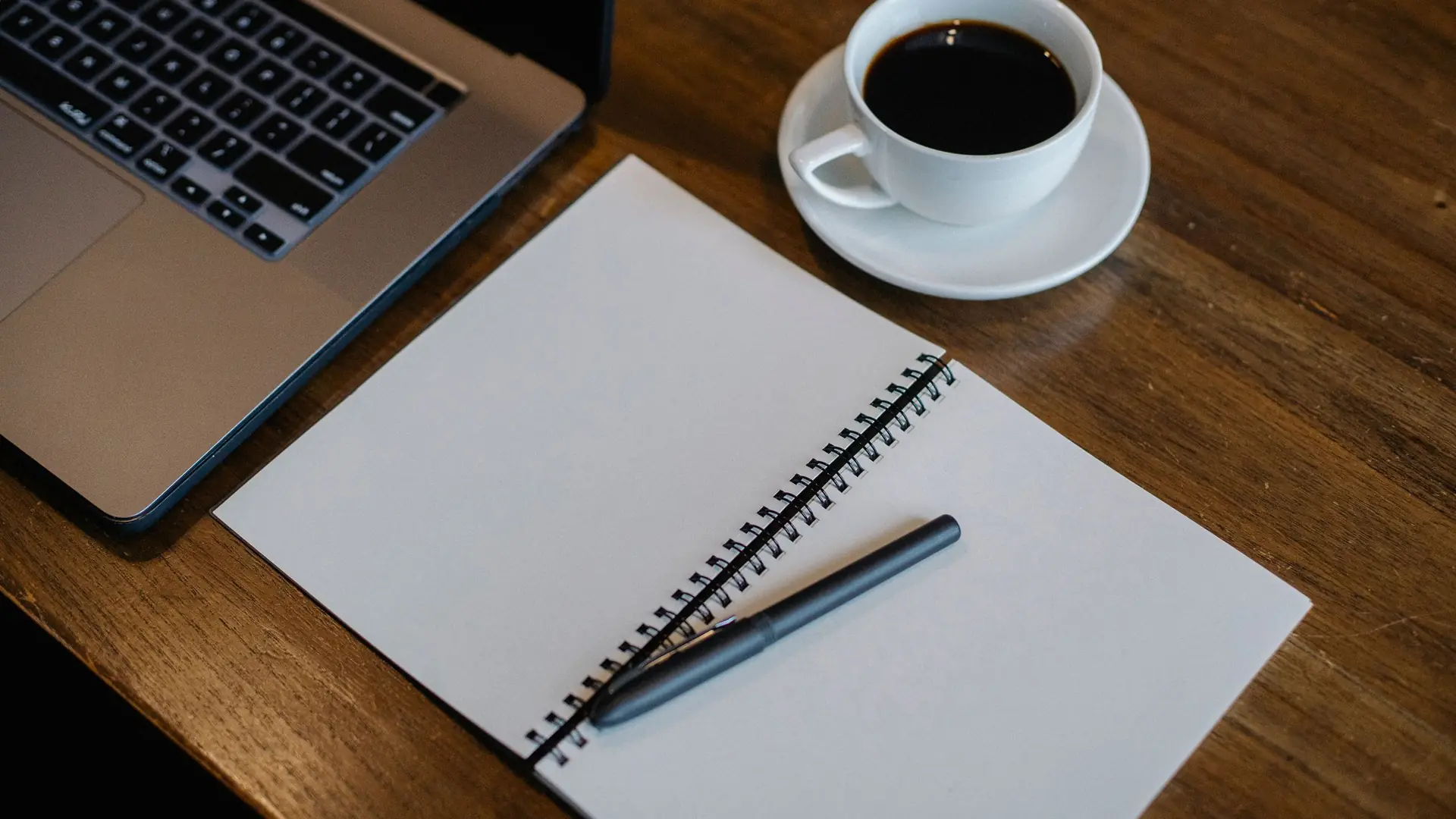 A spiral notebook, a laptop and a cup of coffee on a wooden desk